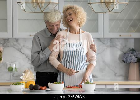 Older husband hugs his cheerful wife preparing vegetarian dish Stock Photo