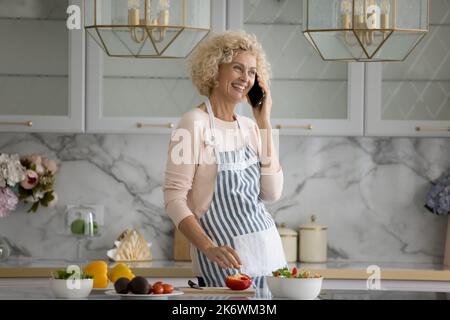 Older woman standing in kitchen talks on smart phone Stock Photo