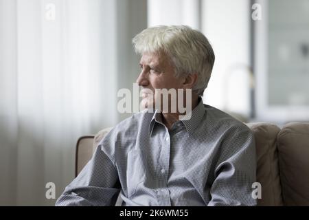 Gloomy elderly man sits on sofa staring into distance Stock Photo