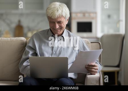 Smiling elderly man sits on sofa using laptop, holding papers Stock Photo