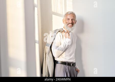 Smiling stylish senior older business man standing at white wall in sunlight. Stock Photo