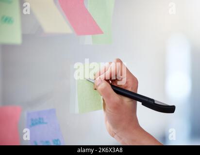Closeup of unknown business woman writing on sticky notes on a transparent board to brainstorm in an office. Caucasian professional sticking a note on Stock Photo