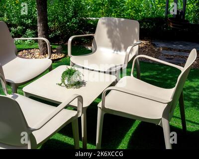 Four empty beige plastic chairs with square shape table with small plant pot in the outdoor garden on artificial grass floor surrounded with the tree. Stock Photo