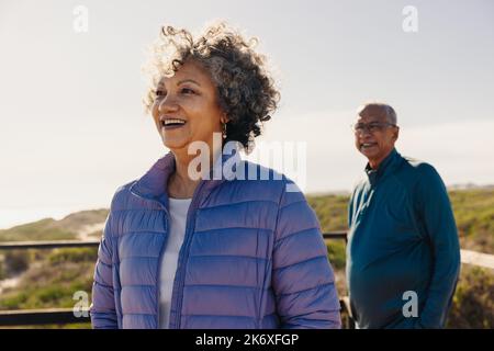 Happy senior woman looking at a refreshing view while standing on a foot bridge with her husband. Cheerful elderly couple enjoying a relaxing seaside Stock Photo