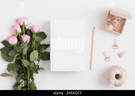 White book mockup with a bouquet of pink roses and workspace accessories on a white table. Stock Photo
