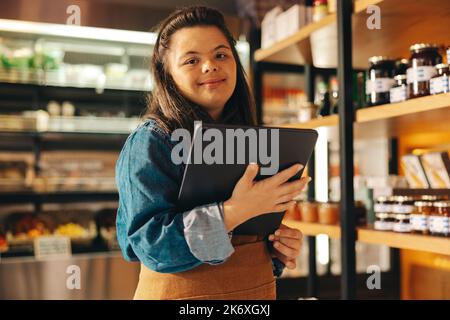 Supermarket employee with Down syndrome using a digital tablet in a shop. Confident woman with an intellectual disability working as a shopkeeper in a Stock Photo