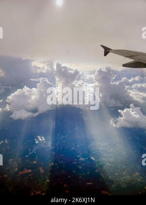Aerial view flying above dramatic fluffy clouds and sun rays piercing shining through the clouds, sunny cloudscape, sunlight, part of airplane, wing, Stock Photo