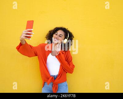 Cheerful african american young woman wearing headphones making peace symbol and taking a selfie over yellow wall Stock Photo