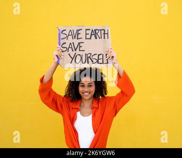Cheerful African American young woman holding a sign that says: Save earth save yourself. Environmentalism, not planet b Stock Photo