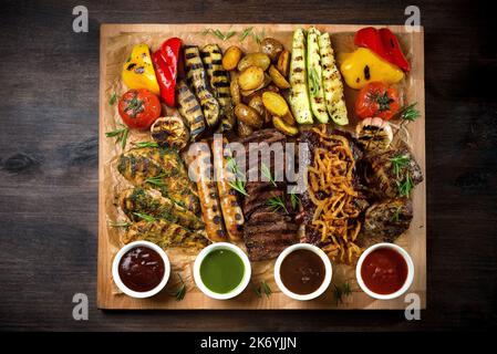 A board with grilled appetizers and sauces. Grilled vegetables and meat snacks: sausages, chicken fillets, pork and beef steaks. Stock Photo