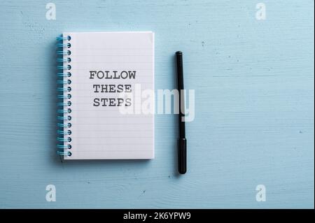 Spiral notebook with a Follow these steps sign placed over pastel blue wooden background with a black marker next to the notepad. With copy space on t Stock Photo