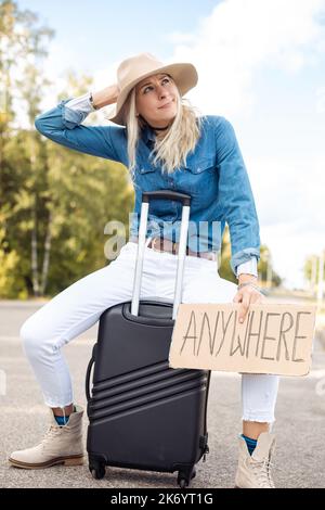 Young thoughtful woman scratching head, holding cardboard with inscription anywhere, sitting on suitcase near forest. Stock Photo