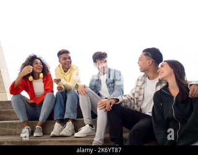 Group of diverse young friends laughing and talking to each other sitting on some stairs in a city Stock Photo