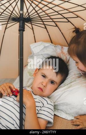 Boy glaring at the camera while lying down in the room under an umbrella next to his sister. Kids party. Fun family. Stock Photo