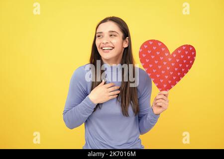 Girl feeling love. Beautiful girl holding valentines gift on yellow background. Portrait of young woman holding red paper heart. Stock Photo