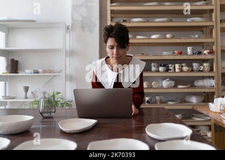 Young pottery entrepreneur sell handmade ceramics online, female artisan using laptop in art studio Stock Photo