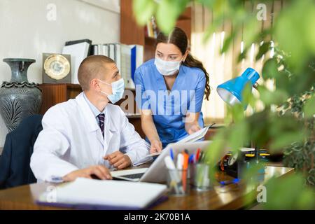 Young doctors colleagues in protective masks discuss an important issue in the office in front of the computer Stock Photo