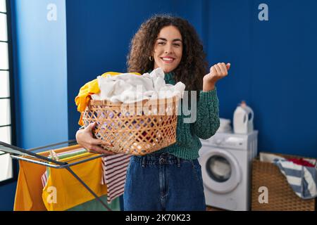 Young hispanic woman holding laundry basket screaming proud, celebrating victory and success very excited with raised arm Stock Photo