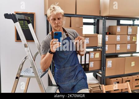 Young blond man using smartphone working at storehouse pointing down looking sad and upset, indicating direction with fingers, unhappy and depressed. Stock Photo