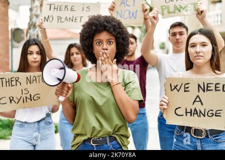 Group of young friends protesting and giving slogans at the street covering mouth with hand, shocked and afraid for mistake. surprised expression Stock Photo