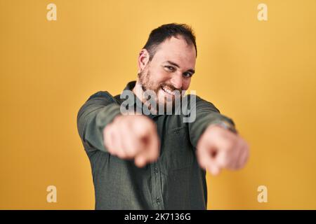 Plus size hispanic man with beard standing over yellow background pointing to you and the camera with fingers, smiling positive and cheerful Stock Photo