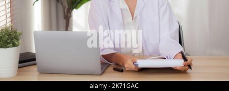 Young woman doctor using laptop computer and looking document paper about patient for diagnostic at clinic, physician female working and reading docum Stock Photo
