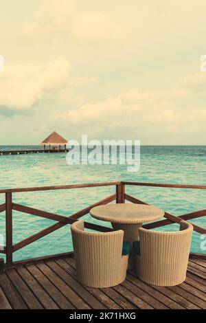 A vertical shot of a wicker table with two armchairs next to it on a wooden floor of a street bar of a luxury resort on a Maldives island with a small Stock Photo