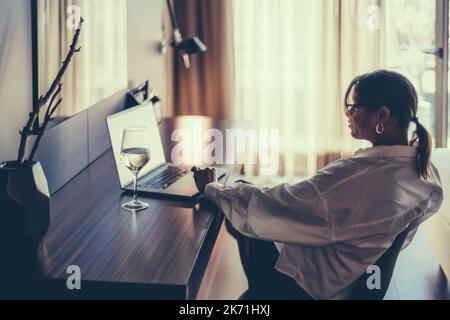 A low-key profile view of a silhouette of a mature Hispanic plus-size female having a video call with her friend via the netbook on the table with a g Stock Photo
