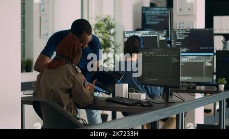 Database developer explaining algorithm to colleague coder pointing at running code on laptop. System engineers collaborating on development group project, using terminal window. Stock Photo