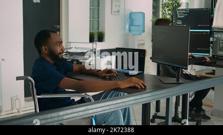 Wheelchair user software engineer working on server program with computer, typing in it coding office. Database programer with chronic disability writing code with system algorithms. Stock Photo
