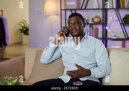 Serious Black man sitting on sofa at home and talking on phone Stock Photo