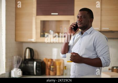 Serious Black man drinking morning coffee and taking on phone with colleague Stock Photo