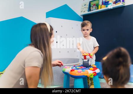 Caucasian little boy standing at the small table in front of others and holding a plastic toy in kindergarten. High quality photo Stock Photo