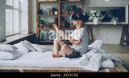 Excited young man in casual clothes is using augmented reality glasses moving hands while sitting on double bed in light apartment. Modern technology and happy people concept. Stock Photo