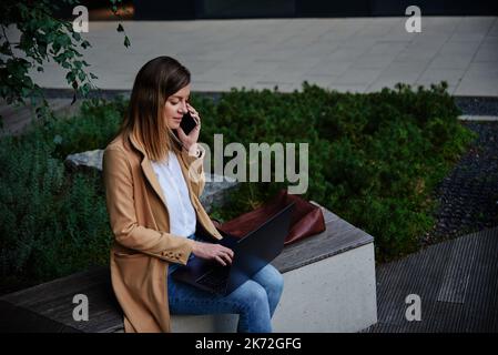 Woman remotely working, sitting on bench at city street and using laptop. Female freelancer work online, typing on laptop and talking with smartphone Stock Photo
