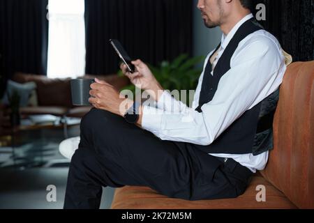 Portrait of a nice and discreet Asian man sitting on a sofa using a smartphone and drinking coffee Stock Photo