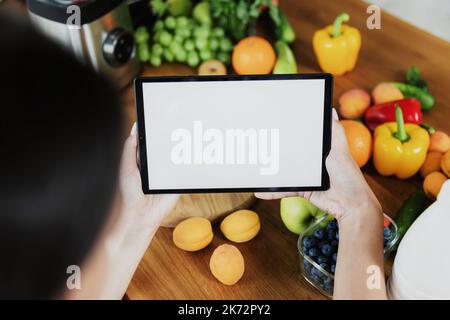 Female hands holding tablet with empty blank white screen mockup in front of vegetables, fruits on table in kitchen at home. Selective focus, cropped. Food delivery concept, mobile diet plan app Stock Photo