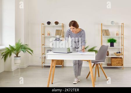 Concentrated busy young woman making notes in notebook while working with laptop in office. Stock Photo