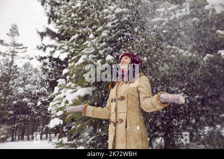 Cheerful young woman having fun and rejoicing in snow falling on her from snow-covered trees. Stock Photo