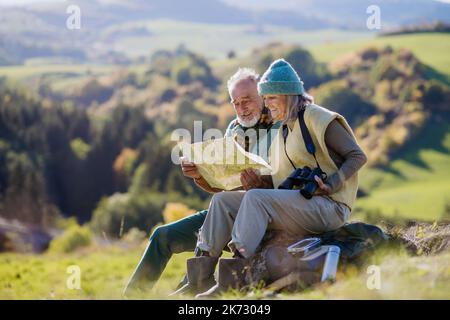 Senior couple having break, looking into paper map during hiking in autumn nature. Stock Photo