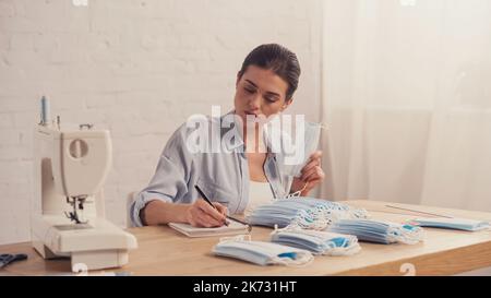 Seamstress writing on notebook near medical masks and blurred sewing ...