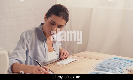 Seamstress writing on notebook near medical masks and blurred sewing ...