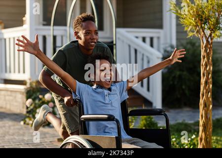 Portrait of black teenage boy pushing littlle brother in wheelchair while having fun together outdoors Stock Photo