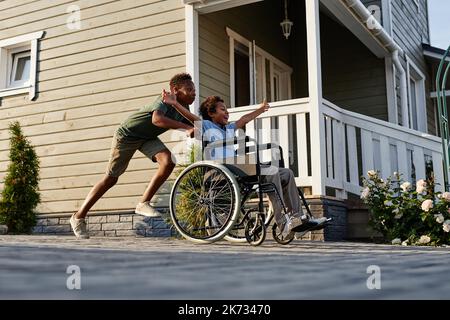 Side view portrait of black teenage boy pushing littlle brother in wheelchair while having fun together outdoors Stock Photo