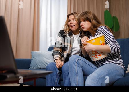 Two scared women of different ages sitting on a sofa with a bucket of popcorn while watching a thriller on a laptop Stock Photo