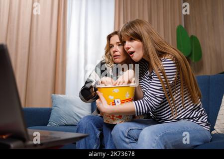 Excited women of different ages watching scary movie and eating popcorn at home on sofa Stock Photo