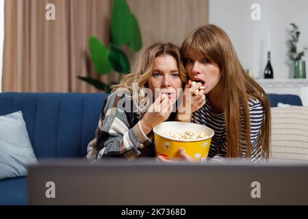 Two women of different ages watch horror movie and eat popcorn while sitting on the couch hugging each other Stock Photo