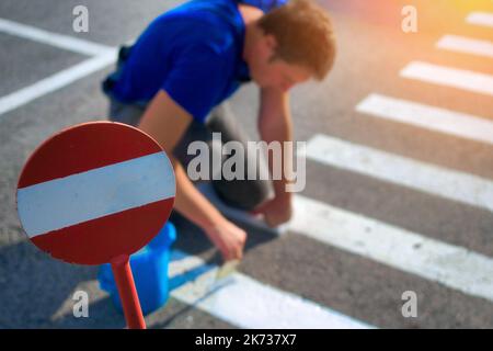 Road service worker paints striped pedestrian crossing on asphalt with paint brush. Markings for pedestrians on roadway. Safe transition for people. Real workflow.. Stock Photo