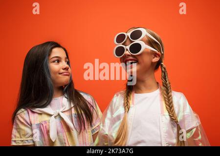 brunette girl looking at cheerful friend having fun and wearing two sunglasses isolated on orange Stock Photo