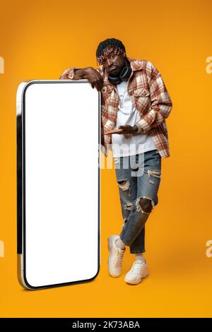 Smiling african man standing near huge smartphone with blank screen on yellow studio background Stock Photo